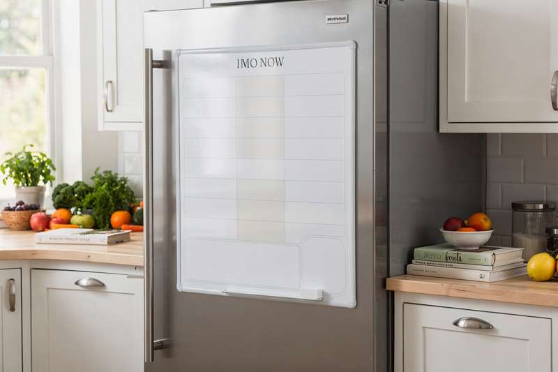 Familie steht zusammen in der Küche und schreibt gemeinsam Mahlzeiten auf ein magnetisches Whiteboard am Kühlschrank.