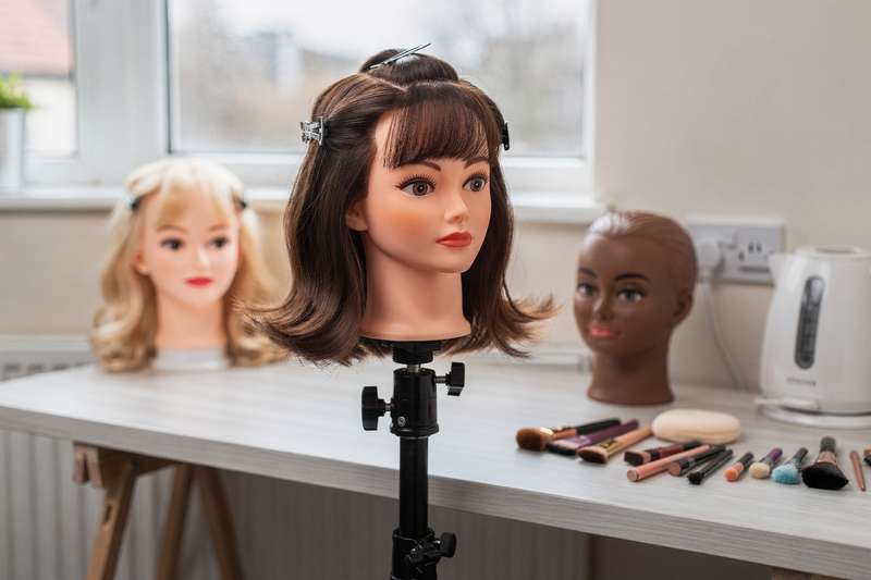 Frisierkopf mit langen blonden Haaren wird von zwei Händen frisiert und mit verschiedenen Styling-Werkzeugen bearbeitet.