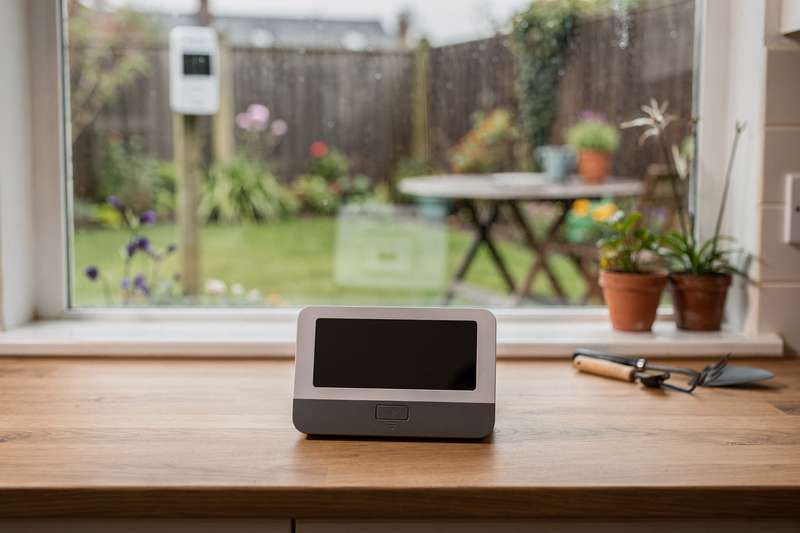 Eine moderne Wetterstation mit digitalem Display steht auf einem Gartentisch neben Blumentöpfen und zeigt Temperatur sowie...